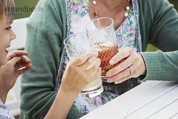 Frau und Junge sitzen im Freien an einem Tisch  halten Getränkegläser in der Hand und klimpern sie zusammen.