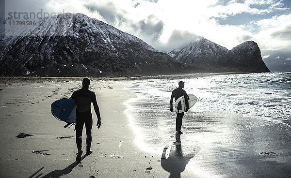 Zwei Surfer in Neoprenanzügen und mit Surfbrettern  die an einem Strand mit Bergen im Rücken entlanglaufen.