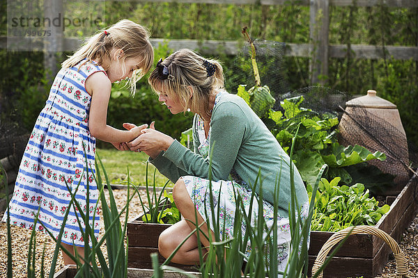 Frau  die in einem Garten an einem erhöhten Gemüsebeet kniet und auf die Hand eines jungen Mädchens neben sich schaut.