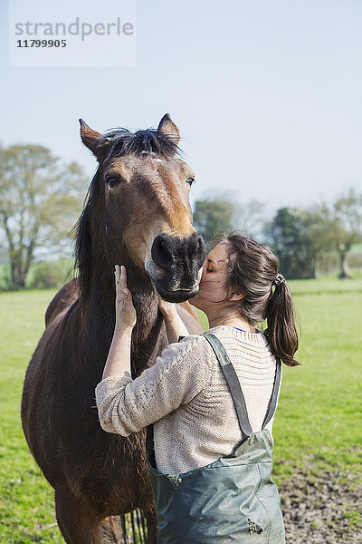 Im Freien stehende Frau  die ein braunes Pferd auf die Wange küsst.