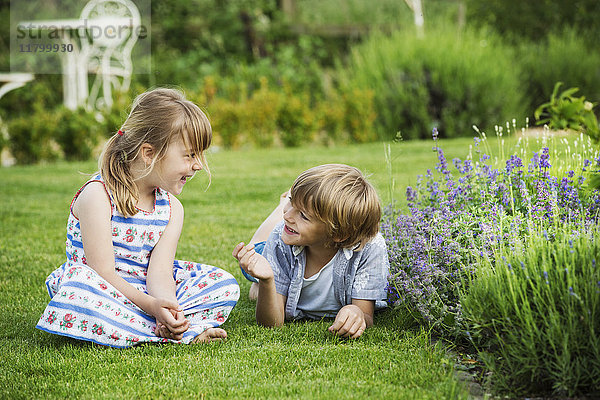 Ein Mädchen sitzt auf dem Rasen und unterhält sich mit ihrem Bruder  der neben ihr auf einem Rasen in einem Garten liegt.