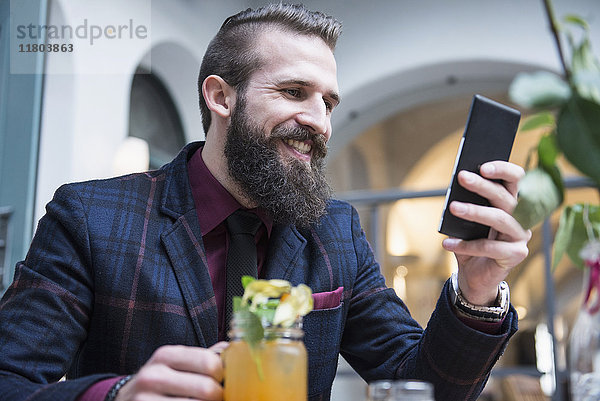 Junger Mann  der ein Mobiltelefon benutzt und ein Mocktail-Glas hält  während er im Restaurant sitzt