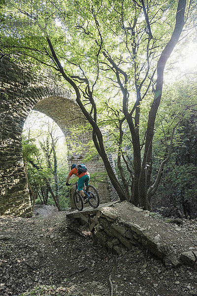 Mann fährt mit dem Fahrrad über einen Fußweg an einer gewölbten Steinmauer im Wald