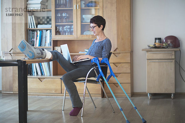 Frau  die ihr gebrochenes Bein auf einem Tisch ausruht und einen Laptop auf einem Stuhl benutzt