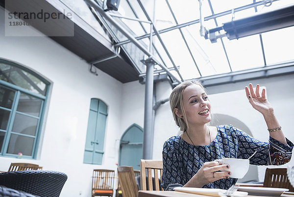 Lächelnde junge Frau winkt und hält eine Kaffeetasse  während sie im Restaurant sitzt