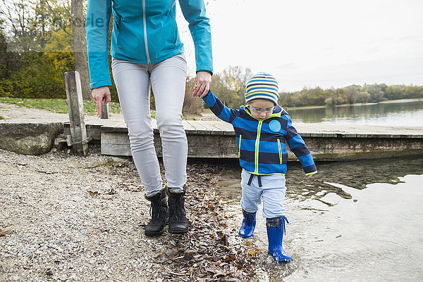 Mutter und Sohn gehen Hand in Hand vor einem Steg am Seeufer im Herbst spazieren