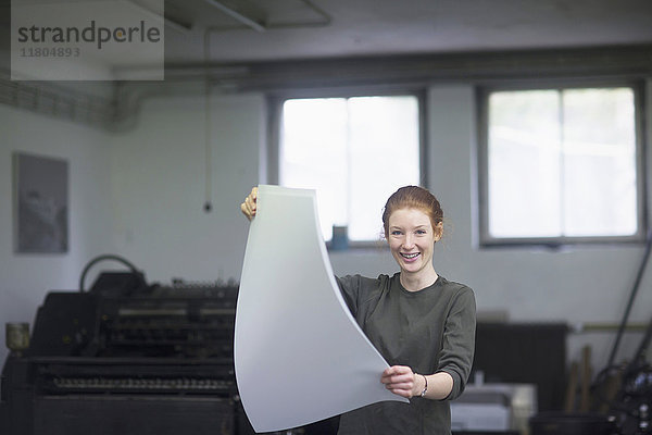 Junge Frau hält ein Bündel Papier an einer Druckerpresse