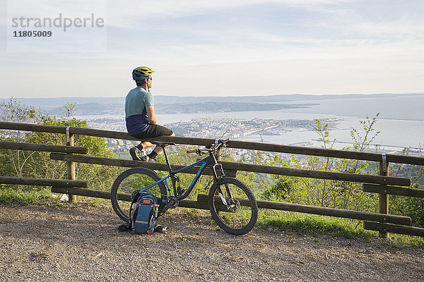 Mountainbiker sitzt auf einem Holzgeländer und schaut auf das Meer und die Stadt