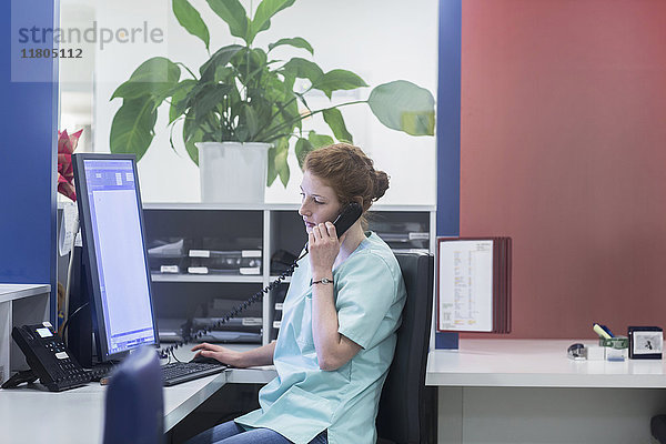 Krankenschwester bei der Arbeit am Schwesternarbeitsplatz  während sie einen Computer benutzt und telefoniert