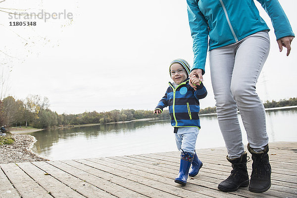 Mutter und Sohn gehen Hand in Hand auf einem Bootssteg über einem See im Herbst