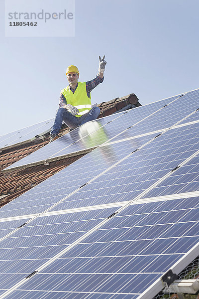 Ingenieur macht eine Friedensgeste  während er eine Pause von der Installation von Solarzellen auf dem Hausdach macht