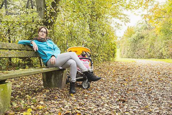 Nachdenkliche Mutter sitzt mit Kinderwagen auf einer Bank im Park