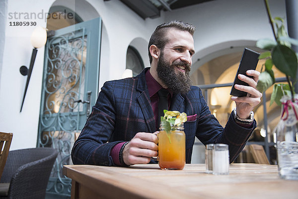 Junger Mann  der ein Mobiltelefon benutzt und ein Mocktail-Glas hält