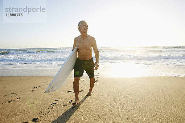 Älterer kaukasischer Mann geht am Strand und trägt ein Surfbrett
