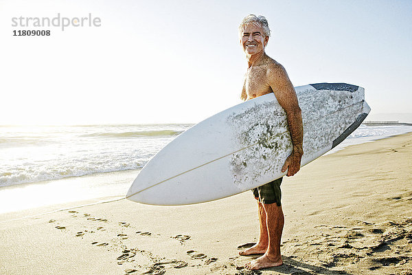 Älterer kaukasischer Mann steht am Strand und trägt ein Surfbrett
