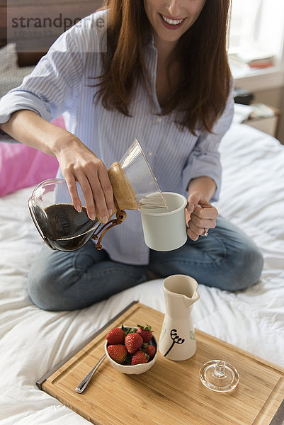 Kaukasische Frau sitzt im Bett und gießt Kaffee ein