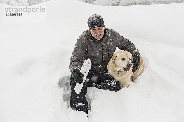 Kaukasischer Mann sitzt im Schnee und umarmt seinen Hund