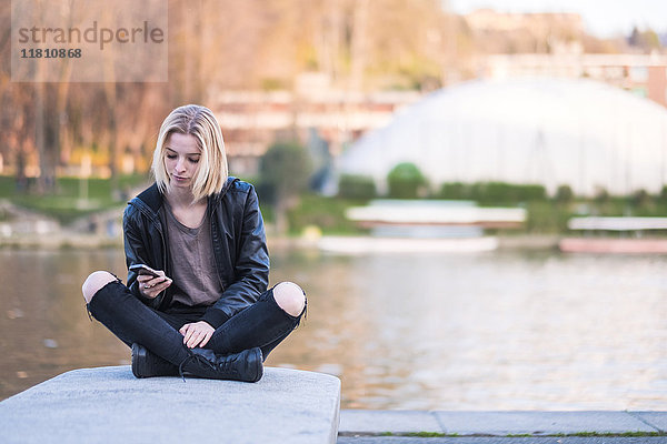 Kaukasische Frau sitzt auf der Mauer in der Nähe des Flusses und schreibt eine SMS auf ihrem Handy