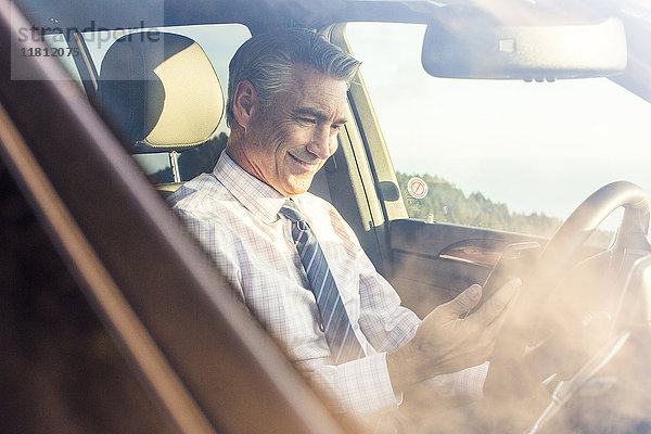 Lächelnder kaukasischer Geschäftsmann sitzt im Auto und schreibt eine SMS auf seinem Handy