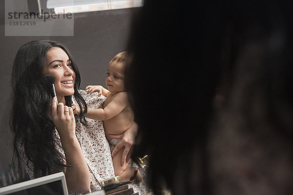 Kaukasische Mutter hält ihren kleinen Sohn und trägt Make-up im Spiegel auf