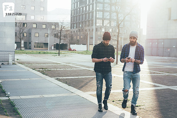 Zwei junge männliche Hipster gehen in der Stadt spazieren und sehen sich dabei Smartphones an