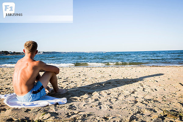 Junger Mann sitzt am Strand und schaut aufs Meer