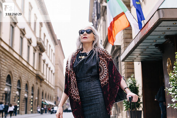 Reife Frau mit langen grauen Haaren mit Rollkoffer vor einem Hotel in Florenz  Italien