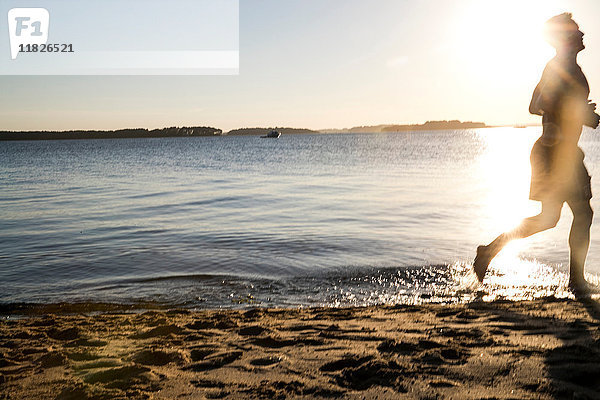 Hinterleuchteter junger männlicher Läufer läuft bei Sonnenuntergang am Strand entlang