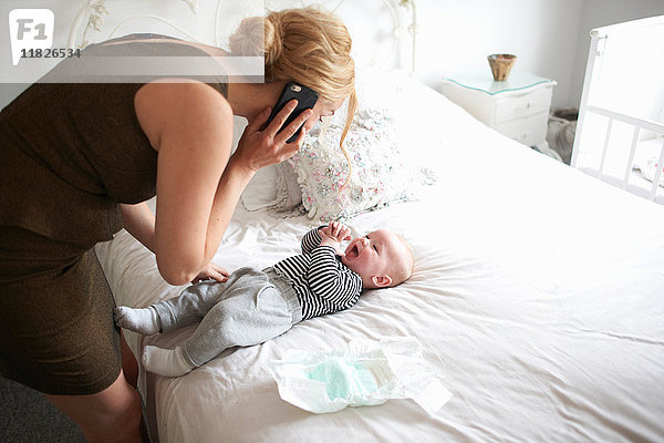 Mutter wechselt die Windel ihres Sohnes während sie ein Mobiltelefon benutzt