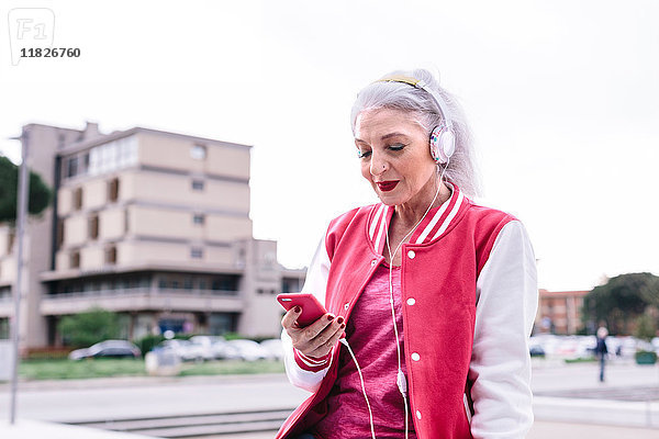 Reife Frau in Baseballjacke  die Kopfhörer hört und ein Smartphone anschaut