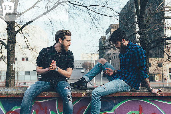 Zwei junge männliche Hipster sitzen an der Wand und schauen auf ein Smartphone