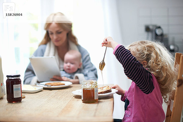 Mutter hält einen kleinen Jungen in der Hand  schaut auf ein digitales Tablett  sitzt mit der kleinen Tochter am Küchentisch  frühstückt