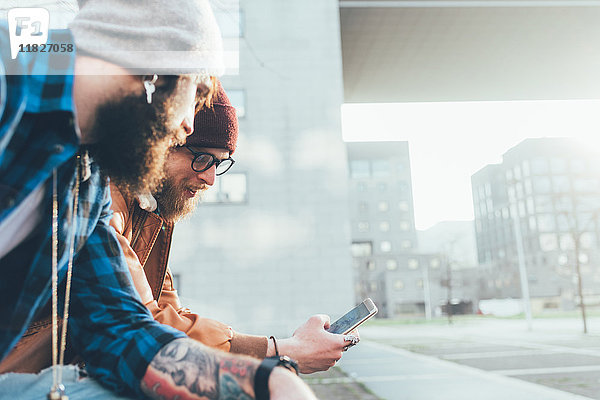 Zwei junge männliche Hipster sitzen an der Wand und schauen auf ein Smartphone  Seitenansicht