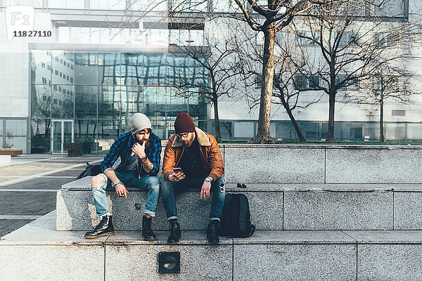 Zwei junge männliche Hipster sitzen an der Wand und schauen auf ein Smartphone