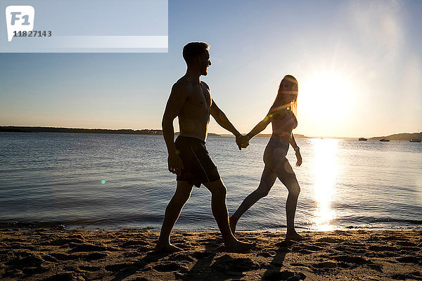 Hinterleuchteter junger Mann und Freundin spazieren Hand in Hand am Strand bei Sonnenuntergang