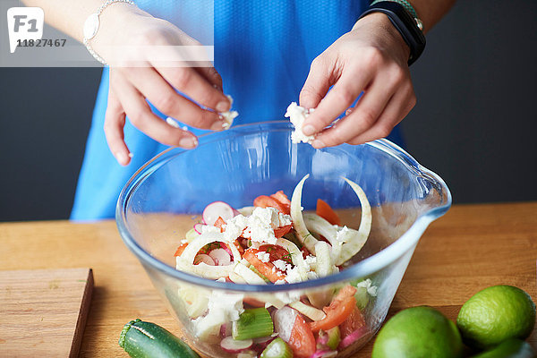 Hände einer jungen Frau am Küchentisch zerbröseln Feta in Salatschüssel