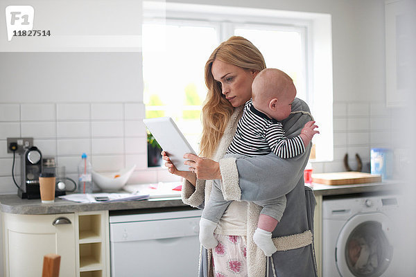Mutter steht in der Küche  hält einen kleinen Jungen und schaut auf ein digitales Tablett