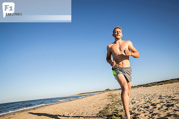 Nacktbrust junger männlicher Läufer läuft am Strand gegen blauen Himmel