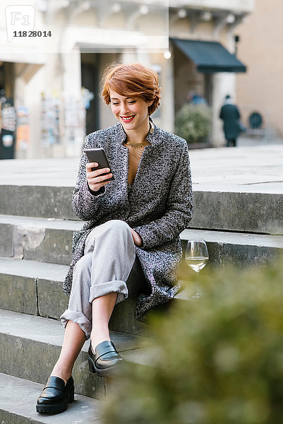 Junge Frau sitzt auf einer Treppe  benutzt ein Smartphone  neben ihr ein Glas Wein
