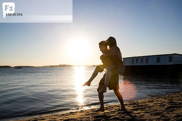 Hinterleuchteter junger Mann gibt seiner Freundin bei Sonnenuntergang Huckepack am Strand