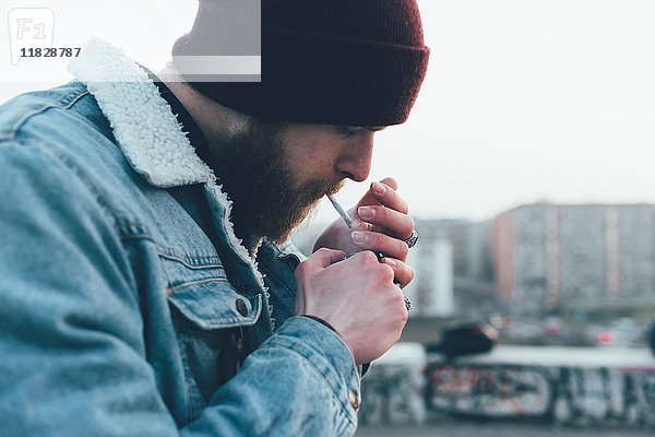 Junger männlicher Hipster mit Strickmütze zündet Zigarette in der Stadt an