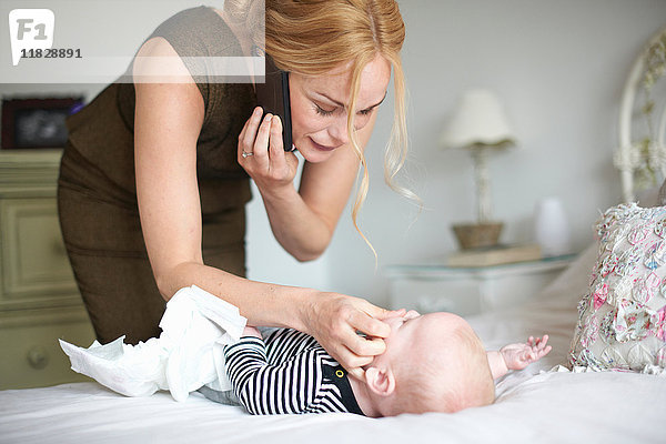 Mutter wechselt die Windel ihres Sohnes während sie ein Mobiltelefon benutzt