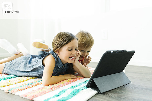 Geschwister (2-3  6-7) liegen auf dem Teppich und schauen auf ein digitales Tablet