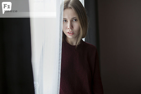 Ernste kaukasische Frau steht in der Nähe eines Vorhangs am Fenster