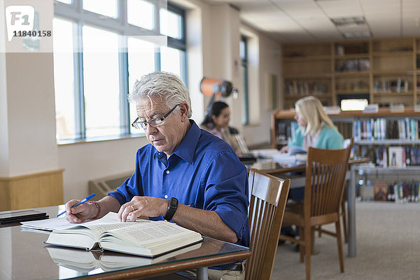 Älterer Mann liest ein Buch in der Bibliothek und schreibt Notizen