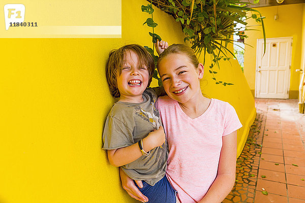 Kaukasisches Mädchen lehnt an einer gelben Wand und hält ihren Bruder