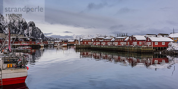 Rote Gebäude säumen die Wasserkante und spiegeln sich im ruhigen Wasser mit Booten  die entlang der Uferlinie vertäut sind; Svolvar  Lofoten  Nordland  Norwegen'.