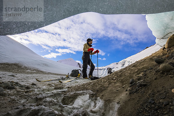 Ein Mann erkundet im Winter auf Skiern die Moräne des Black Rapids Glacier  von einem kleinen Spalt unter dem Eis aus gesehen.