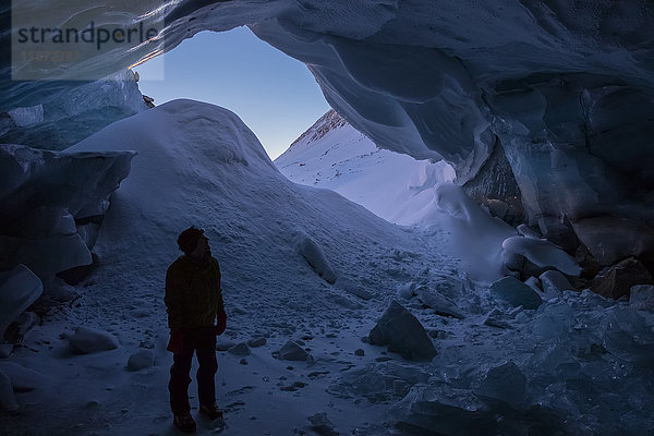 Ein Mann mit Silhouette steht im Eingang einer Eishöhle im Augustana-Gletscher in der Alaska Range im Winter.