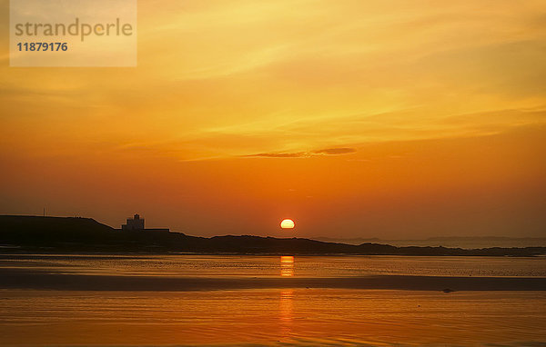 Die goldene Sonne versinkt am Horizont mit einer Silhouette der Küstenlinie und einem orangefarbenen Himmel  der sich im Wasser spiegelt; Bamburgh  Northumberland  England'.